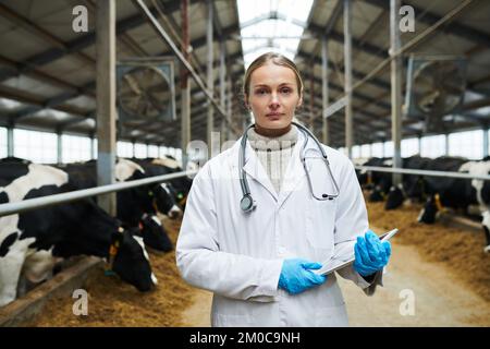 Junge, selbstbewusste Tierärztin in Whitecoat und Handschuhen, die ein digitales Tablet hält und vor langen Kuhställen in die Kamera schaut Stockfoto