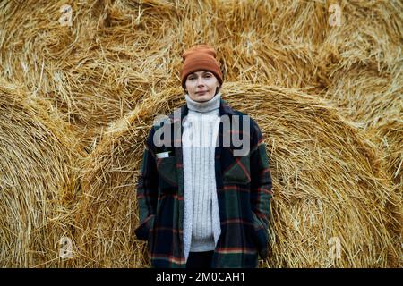 Glückliche junge Arbeiterin einer modernen Farm in Freizeitkleidung, die am riesigen Heuhaufen steht, bestehend aus gerolltem trockenem Stroh, und in die Kamera schaut Stockfoto