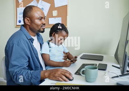 Seitliches Porträt eines lächelnden Vaters mit Tochter, die im Büro zusammenarbeitet, das Kind zur Arbeit mitnimmt Stockfoto