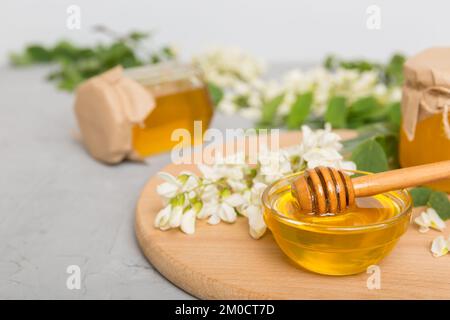 Süßes Honigglas umgab die Akazienblüten des Frühlings. Honig fließt aus einem Löffel in ein Glas. Gläser von klarem frischen Akazienhonig auf Holzgrund. Stockfoto