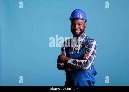 Ein afroamerikanischer Baumeister mit einem fröhlichen Gesicht, der einen Schutzhelm trägt und lächelt, während er in die Kamera schaut. Selbstbewusster Konstrukteur mit gekreuzten Armen auf isoliertem blauem Hintergrund. Stockfoto