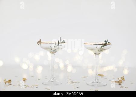 Zwei Trinkgläser mit Champagner und Sekt. Weißer Tisch. Konfetti in Form eines goldenen Sterns. Verschwommener Hintergrund mit Bokeh-Lichtern. Festliches Stillleben Stockfoto