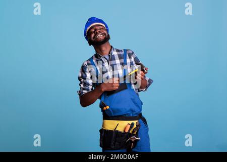 Studiofoto eines frohen afroamerikanischen Baumeisters mit Helm und Gürtelhammer mit zwei Händen. Glücklicher Bauarbeiter, der auf blauem Hintergrund im Studio lächelt. Stockfoto