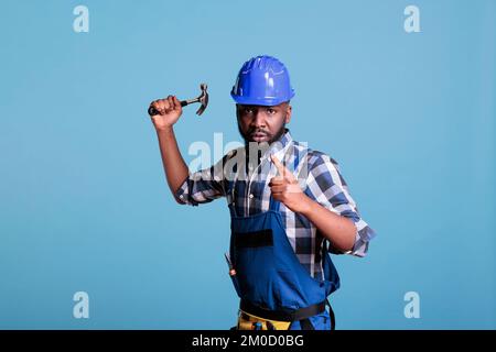 Aggressiver und störender Bauarbeiter, der den Hammer zur Kamera hebt, um zu kämpfen, und bei Studioaufnahmen Gewalt anstiftet. Ein wütender Mitarbeiter mit geballten Fäusten, der eine Show des Kampfes macht. Stockfoto