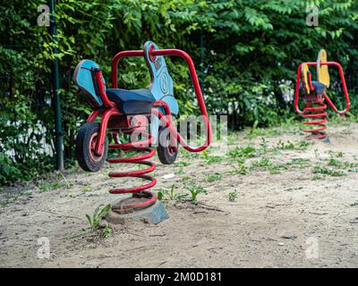 Blaues Motorrad und gelbe Pferdefederschaukel auf einem sandig grünen Vorort Spielplatz Stockfoto