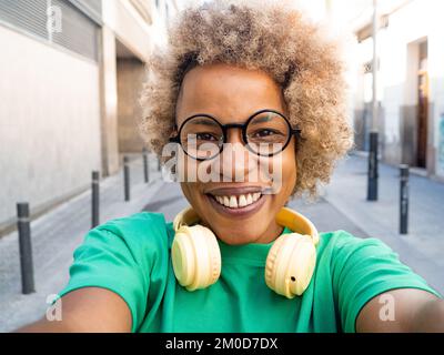Selfie einer glücklichen afroamerikanischen Frau mit Afro-Haaren und Brille und Kopfhörern in der Stadt Stockfoto