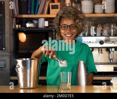 Porträt einer lächelnden afroamerikanischen Barkeeperin mit Afro-Haaren, die in die Kamera schaut, während sie einen Cocktail serviert Stockfoto