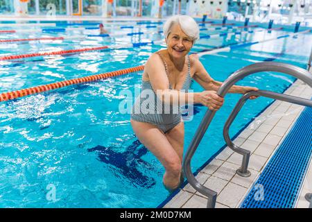 Glückliche, positive, weiße, ältere Frau in einem gestreiften Badeanzug, die Metalltreppen benutzt, um in den Pool zu kommen. Durchgehende Aufnahme in der Halle. Ich schaue in die Kamera. Hochwertiges Foto Stockfoto