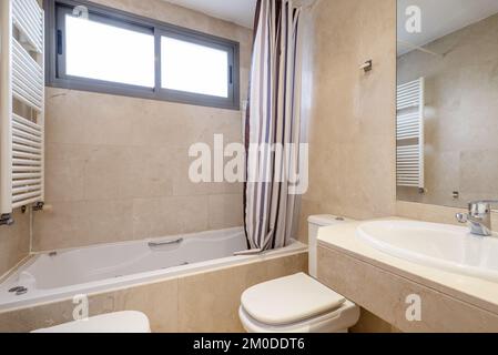 Ein Badezimmer mit cremefarbenen Marmorfliesen, eine Badewanne mit Hydromassagedüsen und ein rahmenloser Spiegel in der Wand Stockfoto