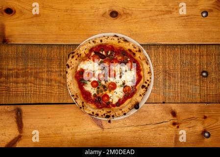 Eine neapolitanische Pizza mit Sardellen, Kapern und Kirschtomaten, Käse und Tomaten, gekocht in einem Steinofen auf einem Holztisch Stockfoto