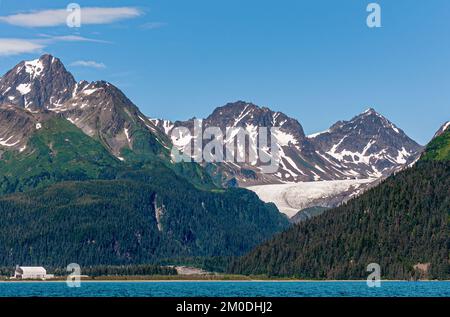 Resurrection Bay, Alaska, USA - 22. Juli 2011: Das weiße Seward-Schiff Trockendock sieht klein aus in der breiten Landschaft der braunen Gebirgszüge mit Schneeflächen und Stockfoto