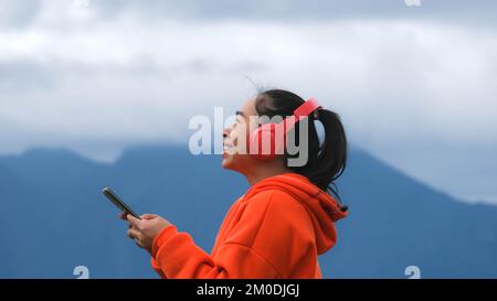 Junge Frau, die auf einem ruhigen Hügel steht und morgens Musik über Kopfhörer hört. Eine Frau, die einen Pullover trägt und die Schönheit der Natur genießt Stockfoto
