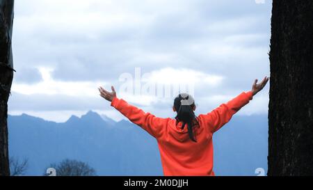 Rückansicht einer jungen Frau, die auf einem ruhigen Berg steht und morgens die Hände auf dem Berg erhebt. Eine Frau, die einen Pullover trägt und die Schönheit genießt Stockfoto