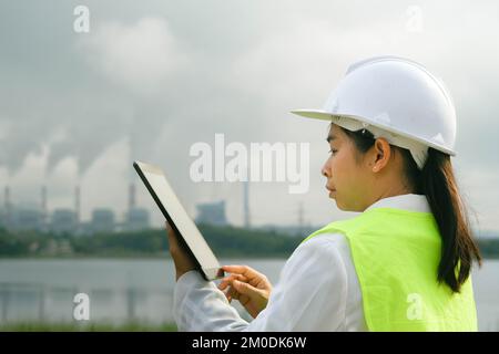 Chefingenieurin mit grüner Weste und Helm steht draußen vor dem Hintergrund eines Kohlekraftwerks und Dampf im Morgennebel. Stockfoto