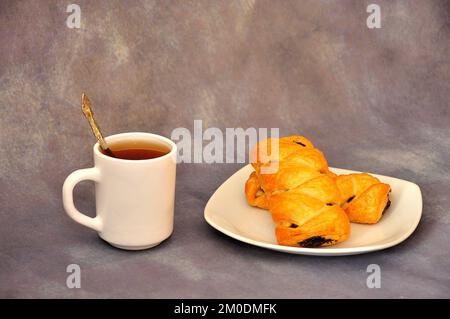 Ein Teller mit zwei Blätterteiggebäck mit Mohnsamen und einer Tasse heißes Getränk auf grauem abstraktem Hintergrund. Nahaufnahme. Stockfoto