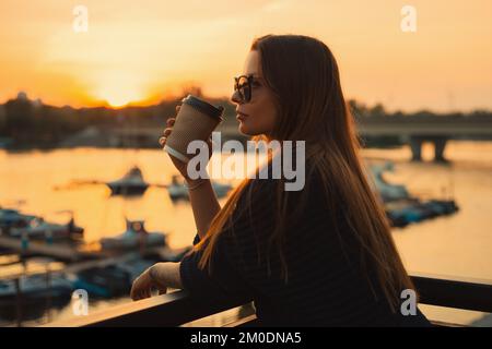 Eine junge, lässige Frau in einer Sonnenbrille, die eine Kaffeetasse hält, lächelt und den Sonnenuntergang am Pier vor dem Fluss im Freien genießt Stockfoto