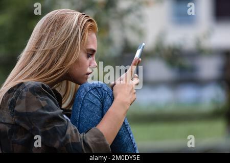 Blondes Mädchen, das sich ein Video auf ihrem Tablet ansieht, sitzt aufgerollt draußen in einem Stadtpark Stockfoto