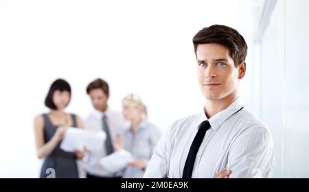 Innovative Teamarbeit führt zum Erfolg. Porträt eines attraktiven Geschäftsführers mit verschränkten Armen - verschwommene Kollegen im Hintergrund. Stockfoto