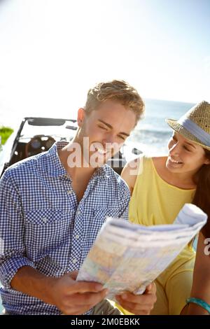 Mal sehen, wie weit wir gekommen sind. Ein junges Paar, das neben seinem Auto am Meer steht und eine Karte liest. Stockfoto