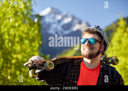 Porträt eines fröhlichen bärtigen Skaters mit Sonnenbrille und Helm, der sein Longboard im Hintergrund im Berg hält Stockfoto