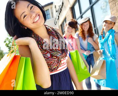 Sie strahlt nach dem Laden. Ein wunderschönes ethnisches Mädchen, das Einkaufstaschen in der Hand hält und mit ihren Freunden im Hintergrund lächelt. Stockfoto