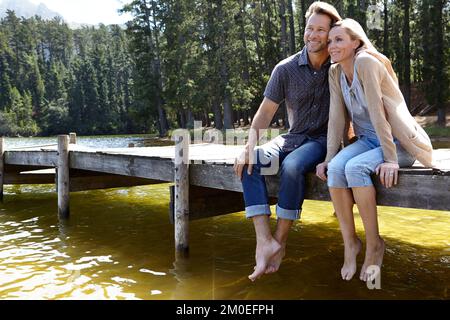 Hingabe am Hafen. Ein liebevolles, reifes Paar, das auf einem Pier auf dem Land an einem See sitzt. Stockfoto