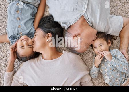 Nahaufnahme einer hispanischen Familie, die zu Hause auf dem Boden lag. Eltern gemischter Rassen küssen ihre beiden Töchter, während sie auf dem liefen. Etage in der Lounge Stockfoto