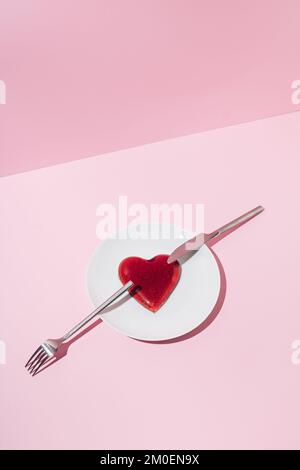 Herz aus rotem Gelee, silbernes Messer und Gabel auf weißem Teller und rosafarbenem Hintergrund. Creative Love oder Valentinstag Minimalkonzept. Stockfoto