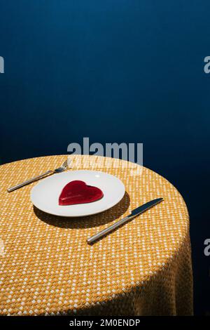 Rotes Gelee-Herz auf weißer Platte und gelbe Tischdecke mit dunkelblauem Hintergrund. Liebe oder Valentinstag Minimalkonzept. Stockfoto