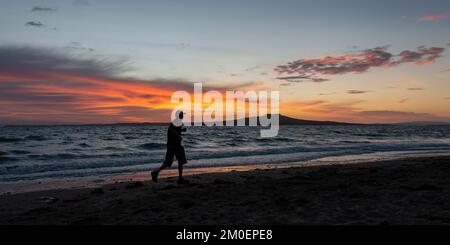 Silhouettenbild eines Mannes, der bei Sonnenaufgang am Milford Beach läuft, Rangitoto Island im Hintergrund, Auckland. Stockfoto