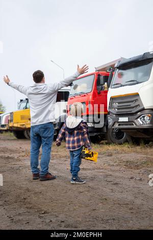 Ein Mann und ein Junge stehen vor großen Lastwagen, Blick von hinten. Sohnes Leidenschaft für Autos, Vater und Kind schauen sich Fahrzeuge auf dem Parkplatz an Stockfoto