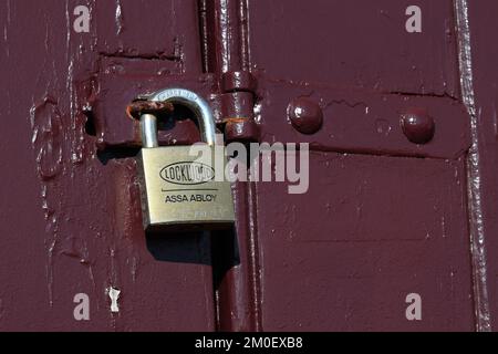 Ein Assa Abloy-Vorhängeschloss mit Lockwood-Emblem an einem kastanienbraunen Türschloss, das eine Holztür verschlossen hält Stockfoto