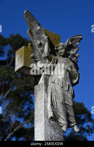 Alte, abgenutzte und verwitterte Steinskulptur eines Engels, der nach oben auf ein heiliges Kreuz oder Kruzifix auf einem Friedhof zeigt Stockfoto