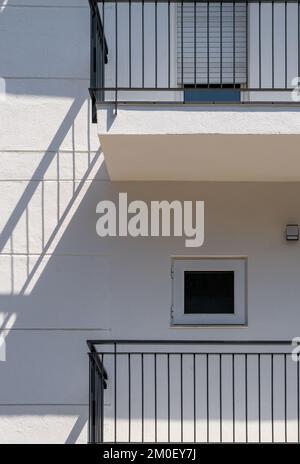 Weiße Fassade eines flachen Gebäudes mit minimalistischem Design und kleinen Fenstern. Der Schatten des schwarzen Geländers wird wie eine Treppe an die Wand projiziert Stockfoto