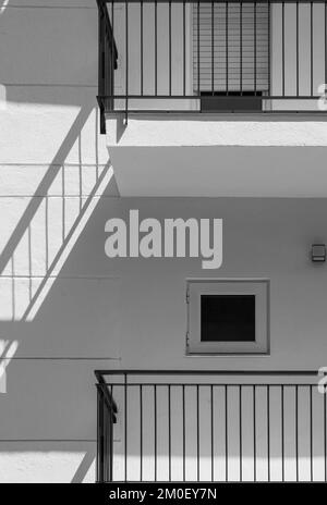 Weiße Fassade eines flachen Gebäudes mit minimalistischem Design und kleinen Fenstern. Der Schatten des schwarzen Geländers wird wie eine Treppe an die Wand projiziert Stockfoto
