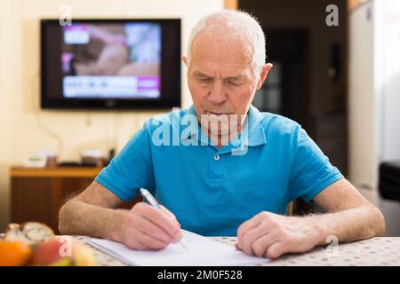 Ein ernster älterer Mann, der im Wohnzimmer einen Brief schreibt Stockfoto