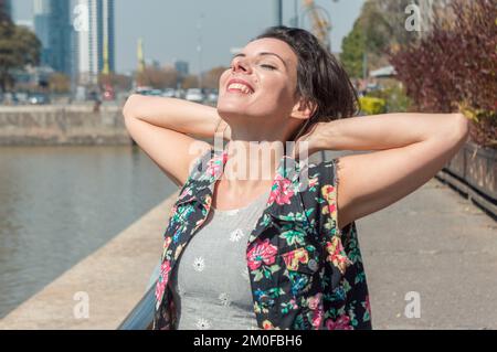 Erwachsene, weiße Touristin mit geschlossenen Augen und lächelnden Augen ist entspannt mit den Händen hinter dem Hals, atmet frische Luft und spaziert durch die Stadt. Stockfoto