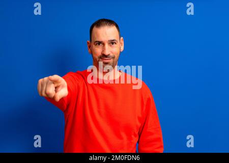 Porträt eines lateinamerikanischen Mannes mit einem Bart, der einen roten Pullover trägt, der nach vorne zeigt und in die Kamera schaut. Porträt halber Länge auf blauem Hintergrund Stockfoto