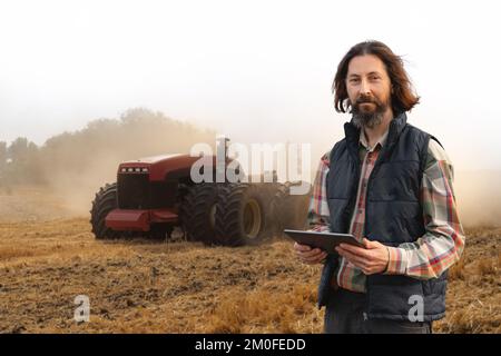 Ein Landwirt mit einem digitalen Tablet steuert einen autonomen Traktor auf einem intelligenten Betrieb Stockfoto