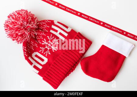 Rotes Band mit Aufschrift Frohe Weihnachten mit rotem Hut, Fäustlingen und roter Socke auf weißem Tisch. Das Konzept der Neujahrsfeier und Stockfoto