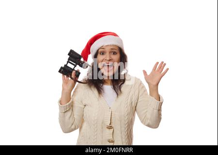 Eine verblüffte Frau mit Weihnachtsmannmütze und Fernglas freut sich auf die bevorstehenden Weihnachtsferien auf weißem Hintergrund Stockfoto