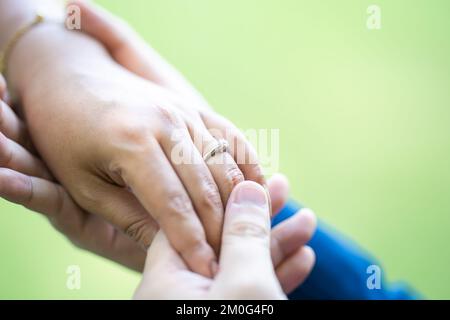 Ein Mann trägt einen Ehering zu einer Frau am linken Ringfinger. Nahaufnahme der Hand auf dem Außenfeld mit Sonnenlicht und Schatten von Bäumen Stockfoto