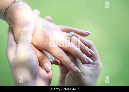 Ein Mann trägt einen Ehering zu einer Frau am linken Ringfinger. Nahaufnahme der Hand auf dem Außenfeld mit Sonnenlicht und Schatten von Bäumen Stockfoto