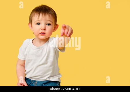 Porträt eines verärgerten Kleinkindes auf Studio-gelbem Hintergrund. Beleidigtes Kind zeigt nach vorne mit Zeigefinger in weißem T-Shirt und blauer Jeans, Kopie Spak Stockfoto