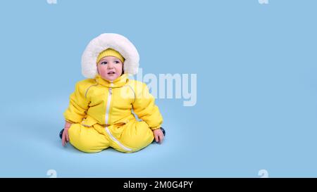 Glückliches Kleinkind in Winterkleidung Schneeanzug auf blauem Studiohintergrund. Ein Kind in einem warmen gelben Overall mit Kapuze. Ein Kind im Alter von einem Jahr, fünf Monaten Stockfoto