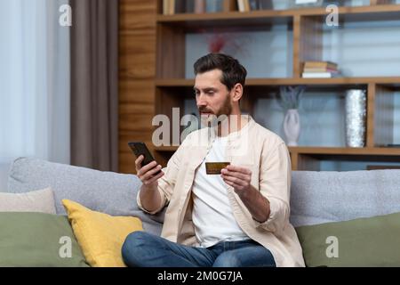 Ein Mann zu Hause sitzt auf dem Sofa, hält eine Bankkreditkarte und ein Mobiltelefon und tätigt Einkäufe in einem Online-Shop. Stockfoto