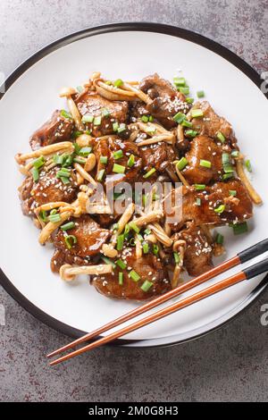 Teriyaki-Rindfleisch mit Shimeji-Pilzen, grünen Zwiebeln und Sesam in Nahaufnahme auf einem Teller auf dem Tisch. Vertikale Draufsicht von oben Stockfoto