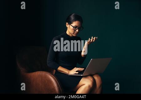 Geschäftsfrau mit Mobiltelefon und Laptop im Sessel. Asiatische Frau, die am Laptop arbeitet und einen Anruf entgegennimmt. Stockfoto