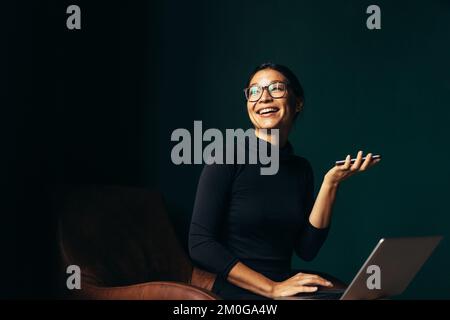 Junge Frau mit Laptop, die über Lautsprecher telefoniert und lächelt. Asiatische Frau, die auf einem Stuhl sitzt, mit Handy und wegsieht. Stockfoto