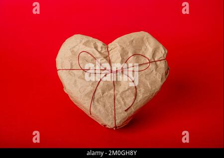 Herzförmige Geschenkbox in Kraftpapier auf rotem Hintergrund Stockfoto